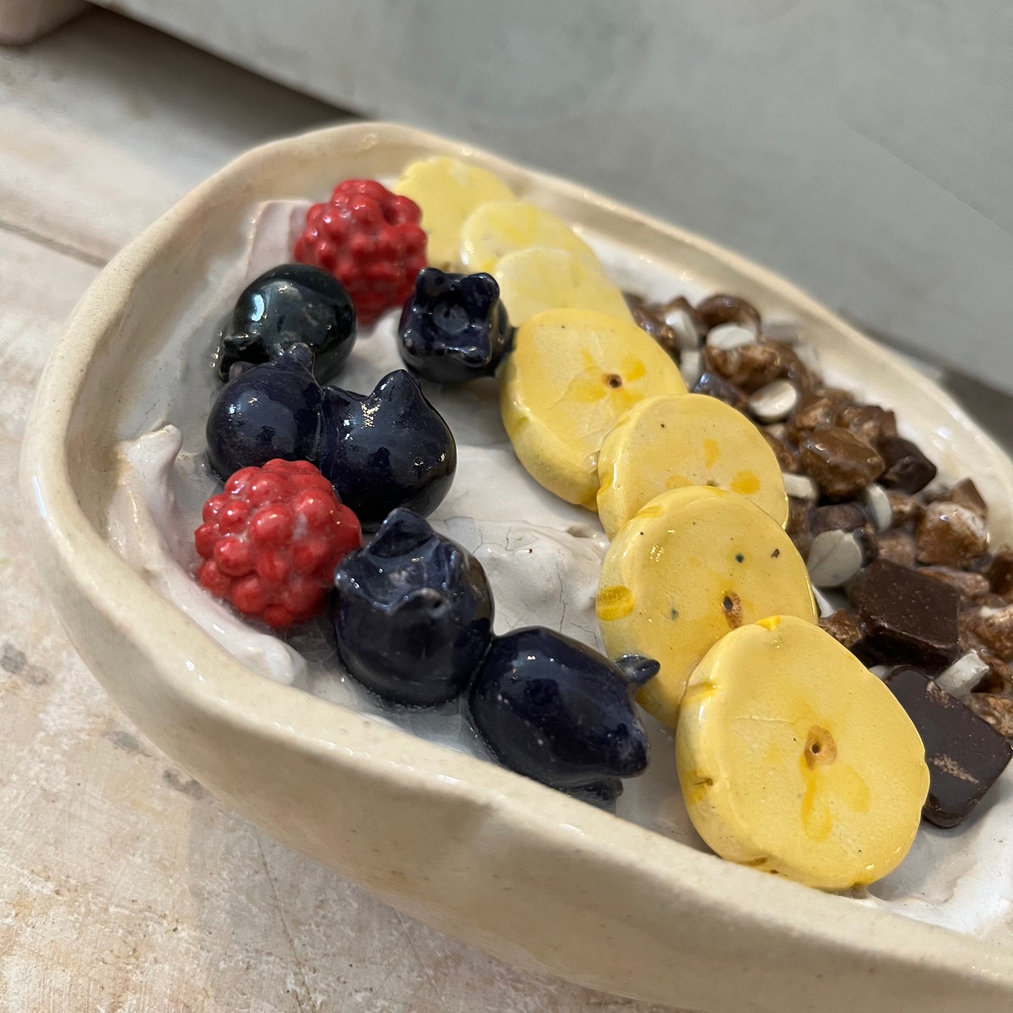 Hyperreal ceramic sculpture of a yogurt bowl with fruit and muesli, handmade in pottery with detailed raspberries, banana, blueberries, and chocolate