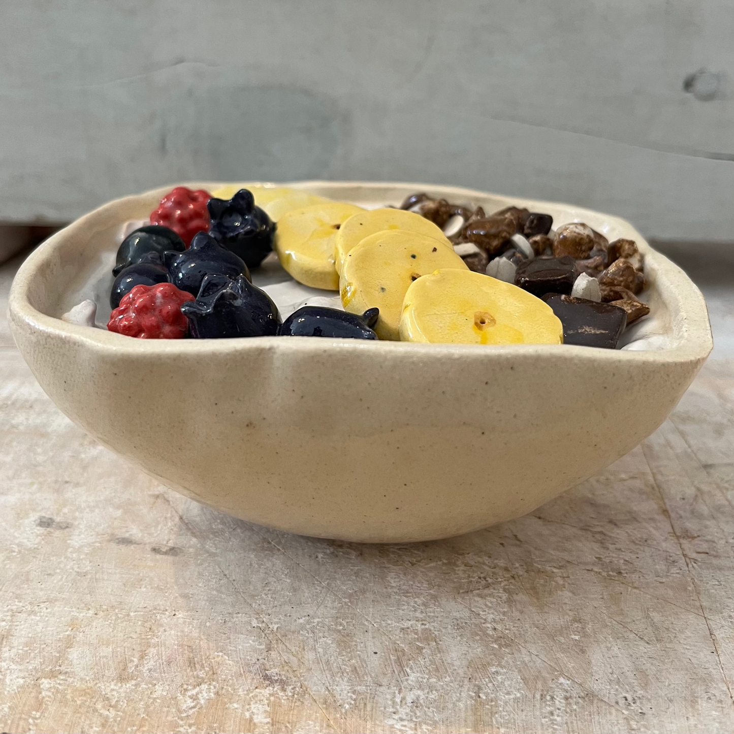 Hyperreal ceramic sculpture of a yogurt bowl with fruit and muesli, handmade in pottery with detailed raspberries, banana, blueberries, and chocolate