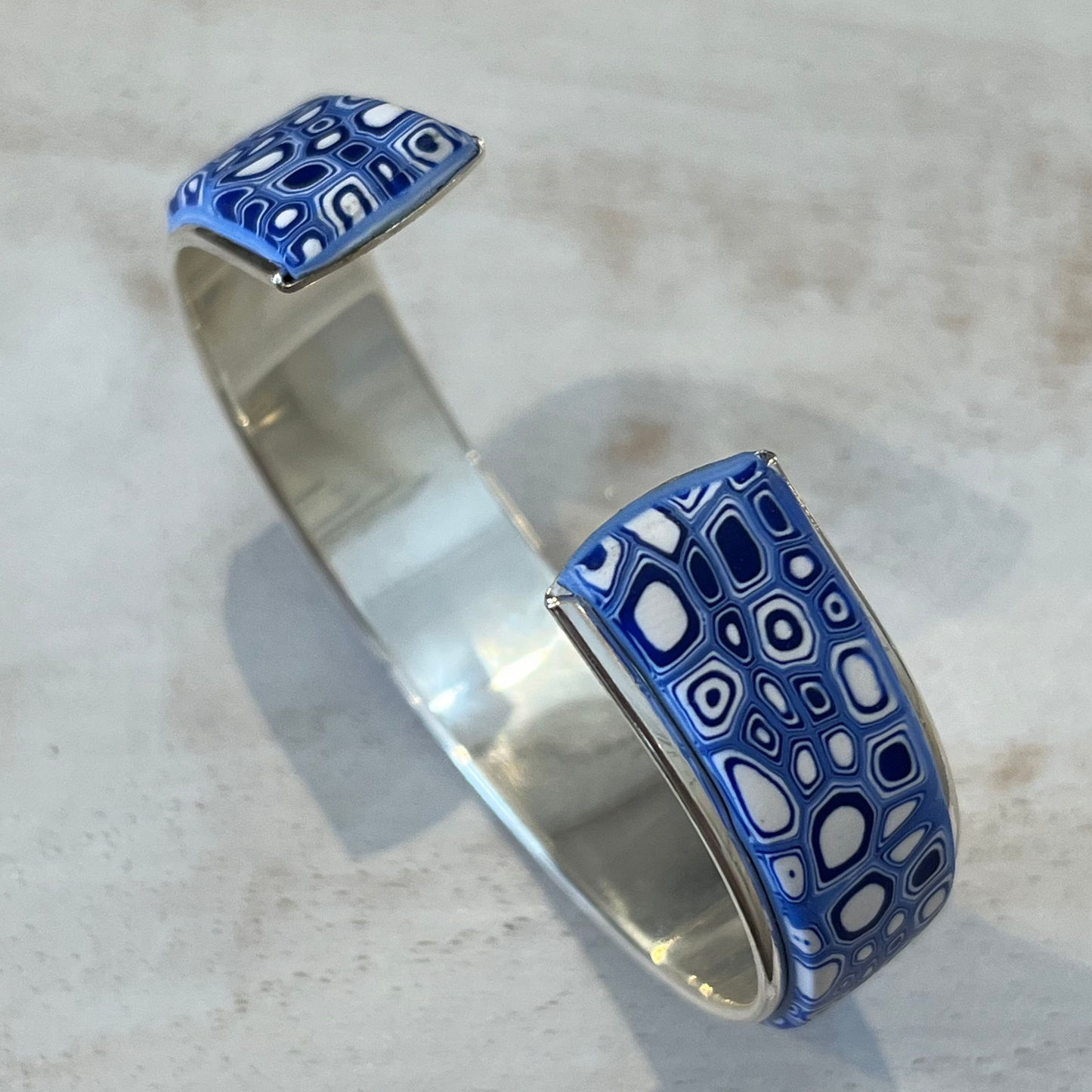 A handcrafted adjustable cuff from Stephanie Fraselle’s Ocean Shimmer collection, featuring an intricate mosaic of tiny, unique cells in sapphire blue, navy, ultramarine, and white. The polymer clay design is set in Sterling Silver 925 over brass, 15 mm wide, 16.3cm long, lead-, cadmium-, and nickel-free.
