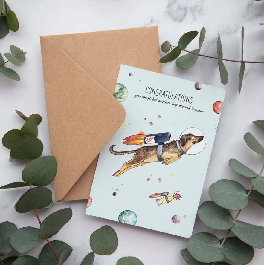 Bamboo greeting card featuring Lottie the dog floating in space with the message “Congratulations, you have completed another trip around the sun”