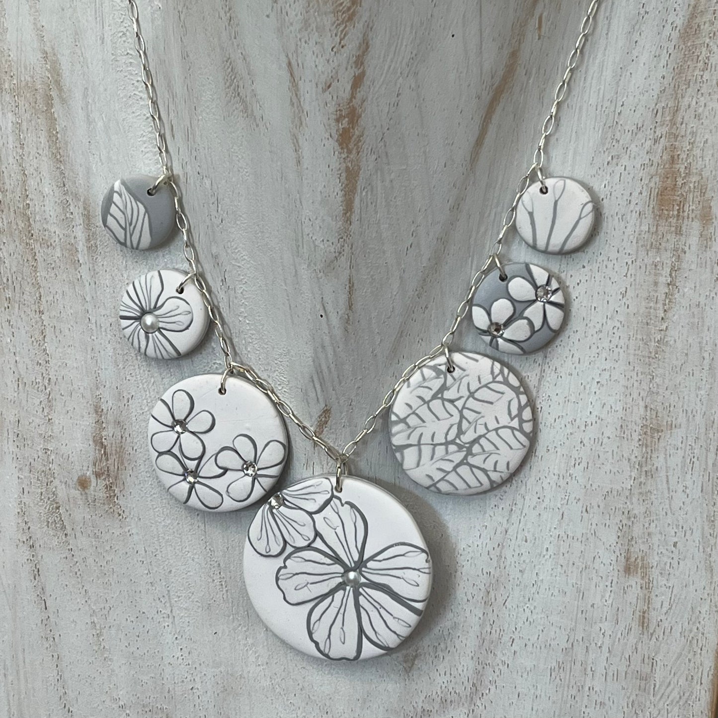 The Seven necklace from the White Blossom Collection by Stephanie Fraselle featuring seven handcrafted polymer clay medallions with botanical motifs, Swarovski crystals, and pearls on a sterling silver chain.