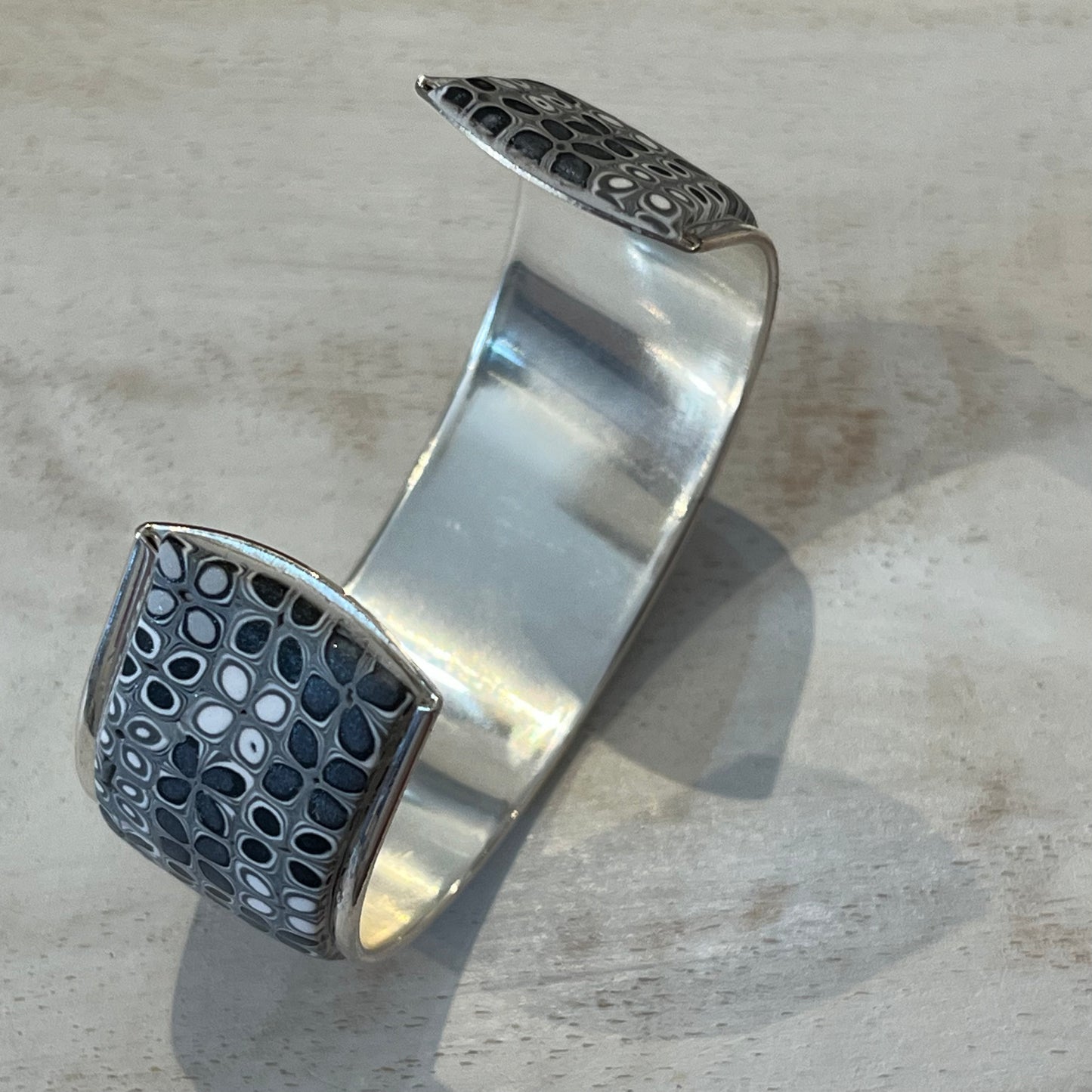 Moonlit Shimmer Cuff by Stephanie Fraselle featuring a hand-placed mosaic of polymer clay tiles in grey, white, and silver tones, set in sterling silver-coated brass.