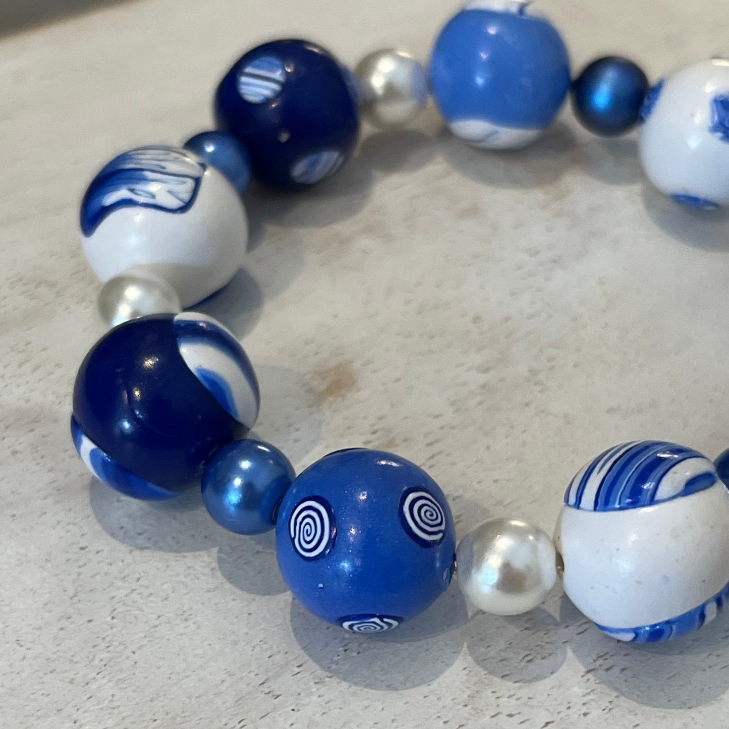 Handcrafted nine-bead bracelet from Stephanie Fraselle’s Marégraphie collection, featuring individually sculpted polymer clay beads in ocean blues and whites, accented with Swarovski crystals and pearls, strung on clear elastic cord, 20 cm length, 15 mm beads, lead-, cadmium-, and nickel-free.