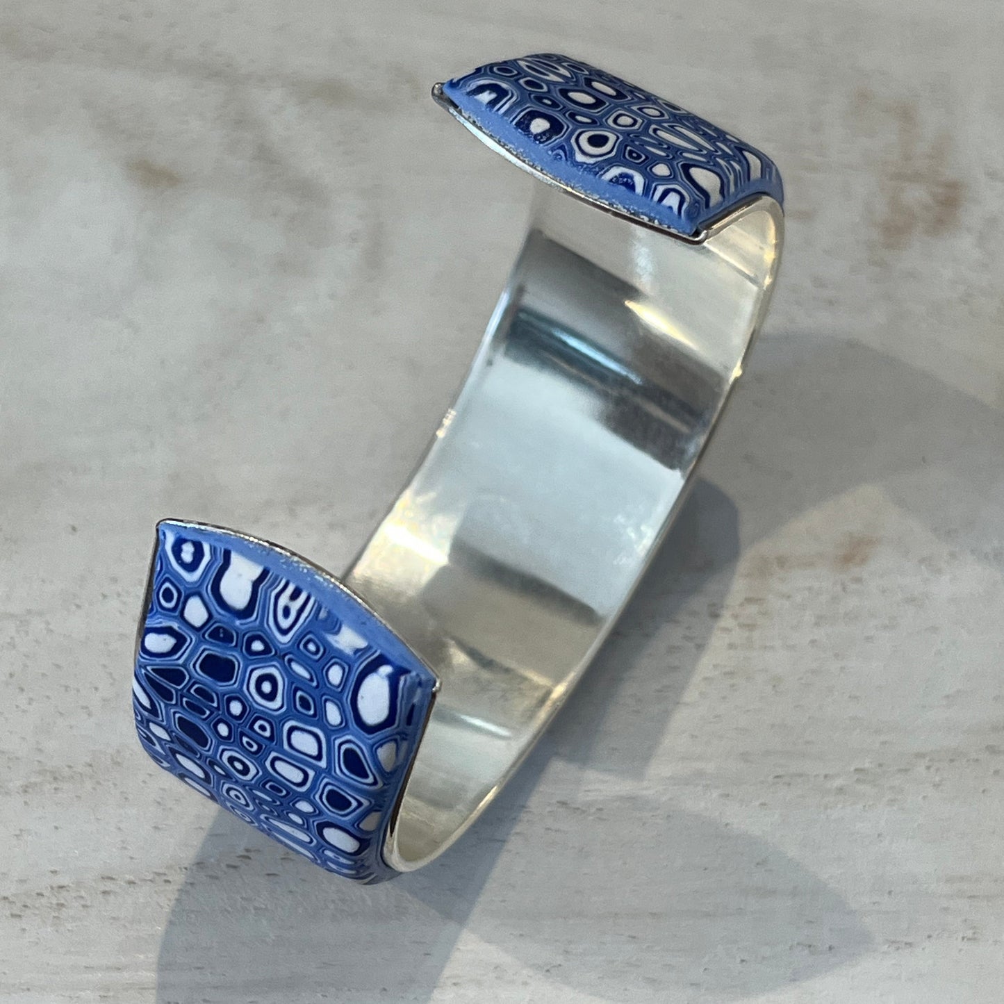 A handcrafted adjustable cuff from Stephanie Fraselle’s Ocean Shimmer collection, featuring an intricate mosaic of tiny, unique cells in sapphire blue, navy, ultramarine, and white. The polymer clay design is set in Sterling Silver 925 over brass, 23 mm wide, 14.5 cm long, lead-, cadmium-, and nickel-free.