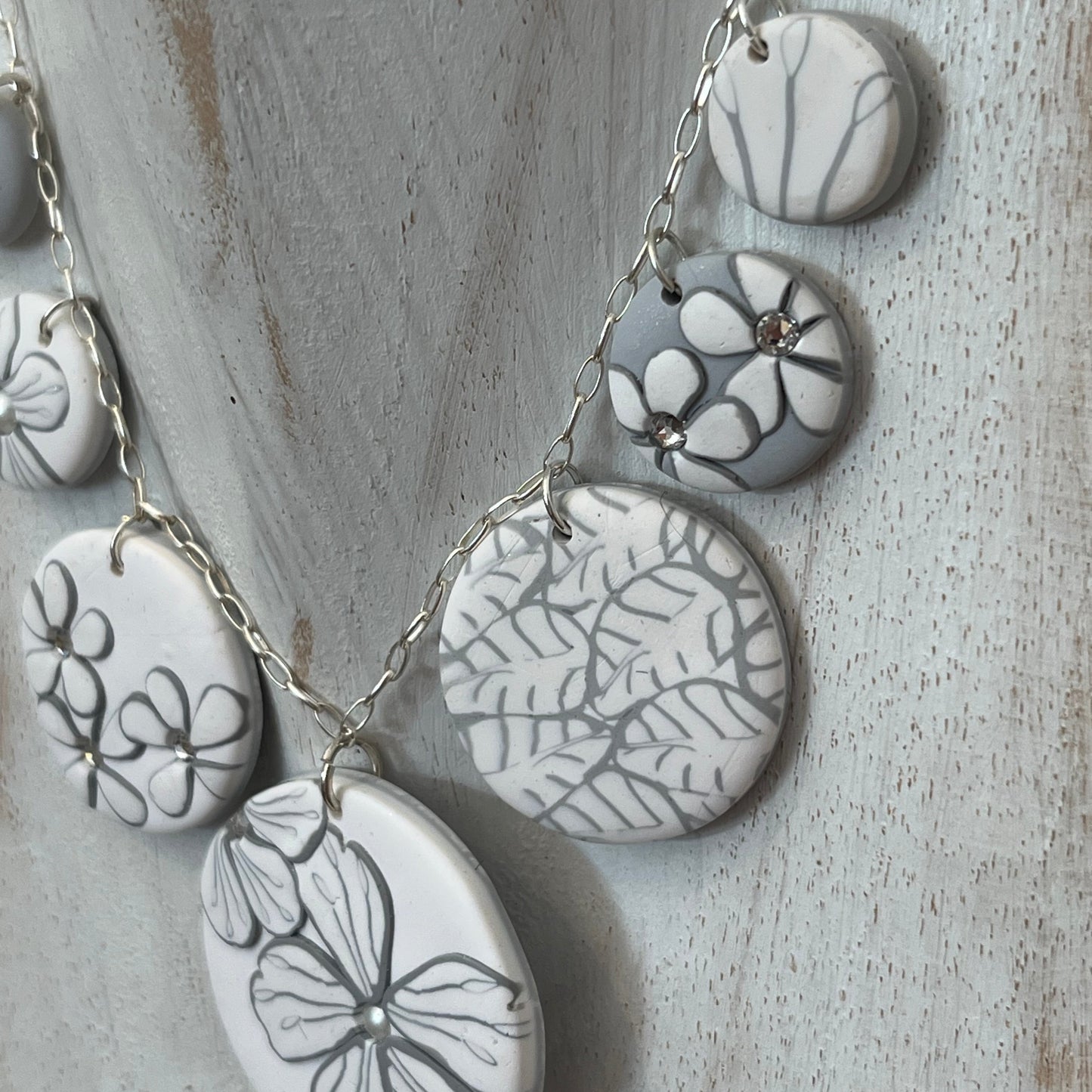 The Seven necklace from the White Blossom Collection by Stephanie Fraselle featuring seven handcrafted polymer clay medallions with botanical motifs, Swarovski crystals, and pearls on a sterling silver chain.