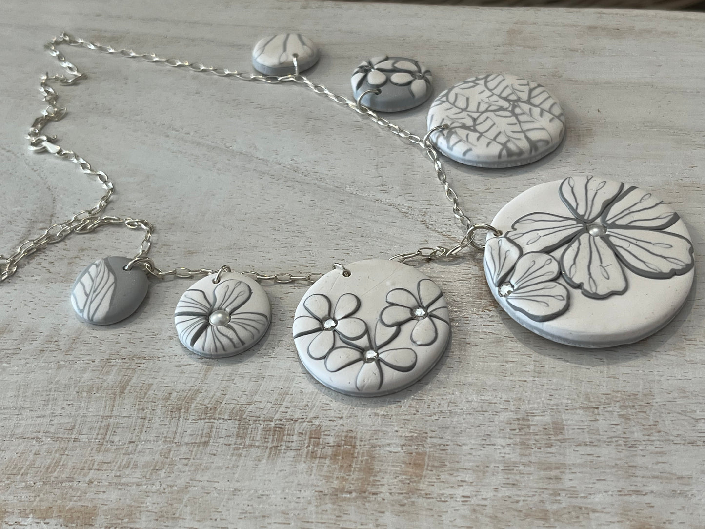 The Seven necklace from the White Blossom Collection by Stephanie Fraselle featuring seven handcrafted polymer clay medallions with botanical motifs, Swarovski crystals, and pearls on a sterling silver chain.