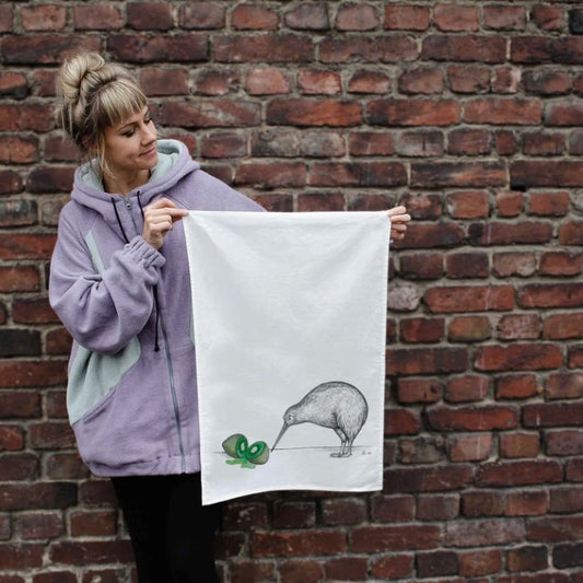 Black and white kiwi bird illustration looking at a colourful watercolour of a sliced green kiwi fruit on a linen tea towel
