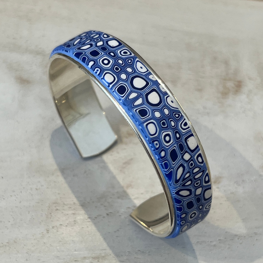 A handcrafted adjustable cuff from Stephanie Fraselle’s Ocean Shimmer collection, featuring an intricate mosaic of tiny, unique cells in sapphire blue, navy, ultramarine, and white. The polymer clay design is set in Sterling Silver 925 over brass, 15 mm wide, 16.3cm long, lead-, cadmium-, and nickel-free.