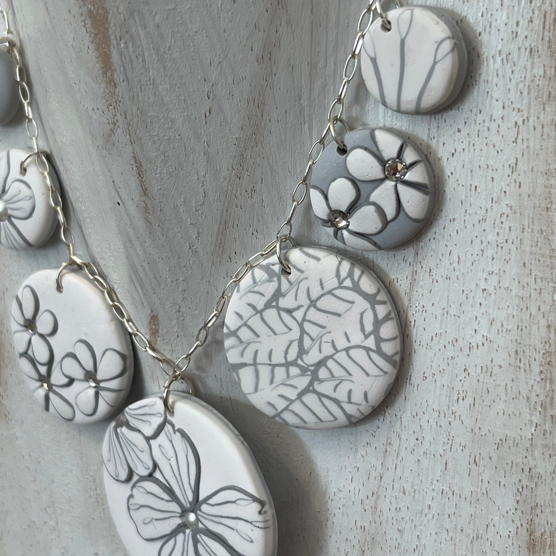 The Seven necklace from the White Blossom Collection by Stephanie Fraselle featuring seven handcrafted polymer clay medallions with botanical motifs, Swarovski crystals, and pearls on a sterling silver chain.